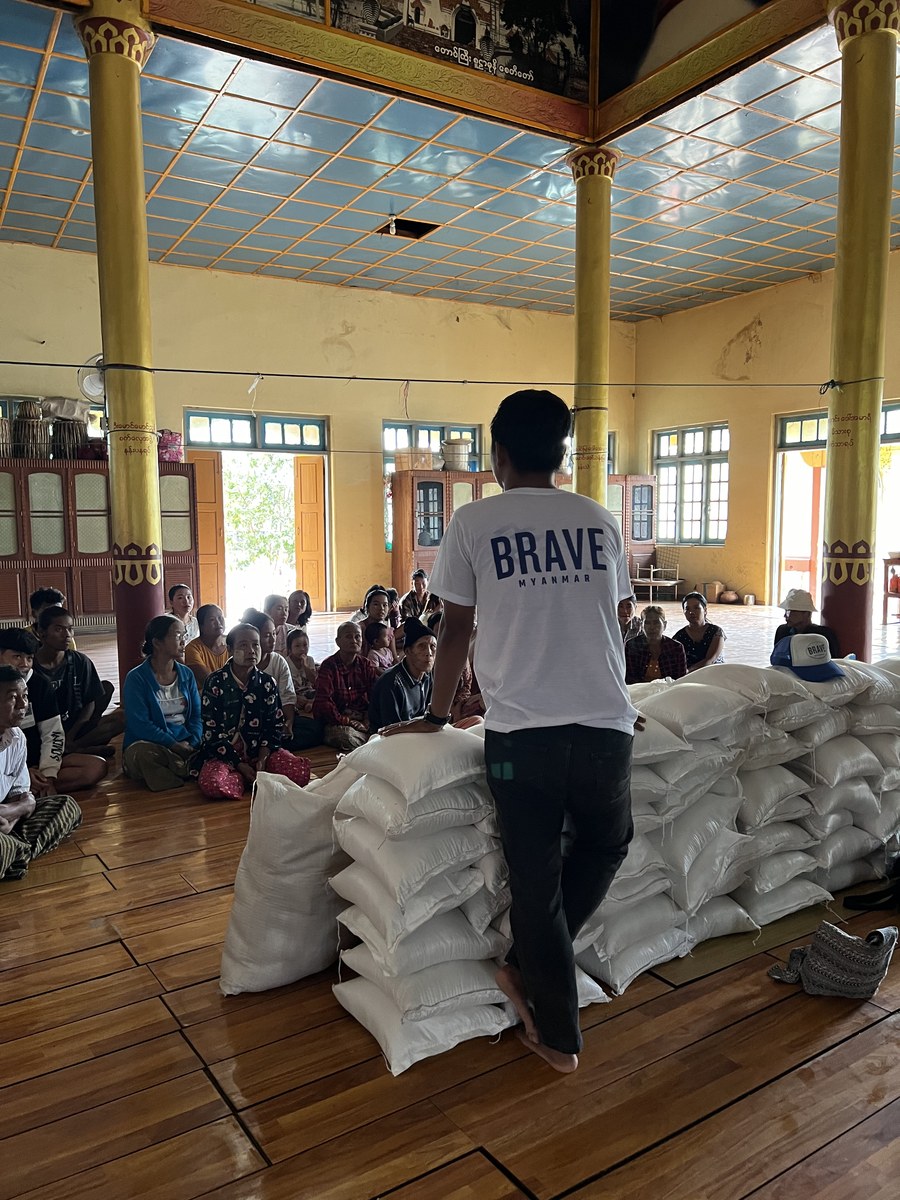 BRAVE volunteer addressing a seated crowd from atop stacked rice bags in a temple hall